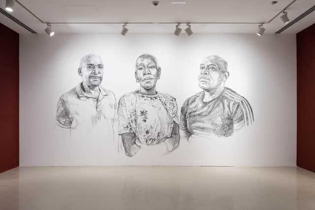 The exhibition features large-scale charcoal portraits drawn directly on to the gallery wall and eight framed works on paper of people impacted by the scandal layered over hand-drawn reproductions of identity papers, evidence of their right to remain in the UK.