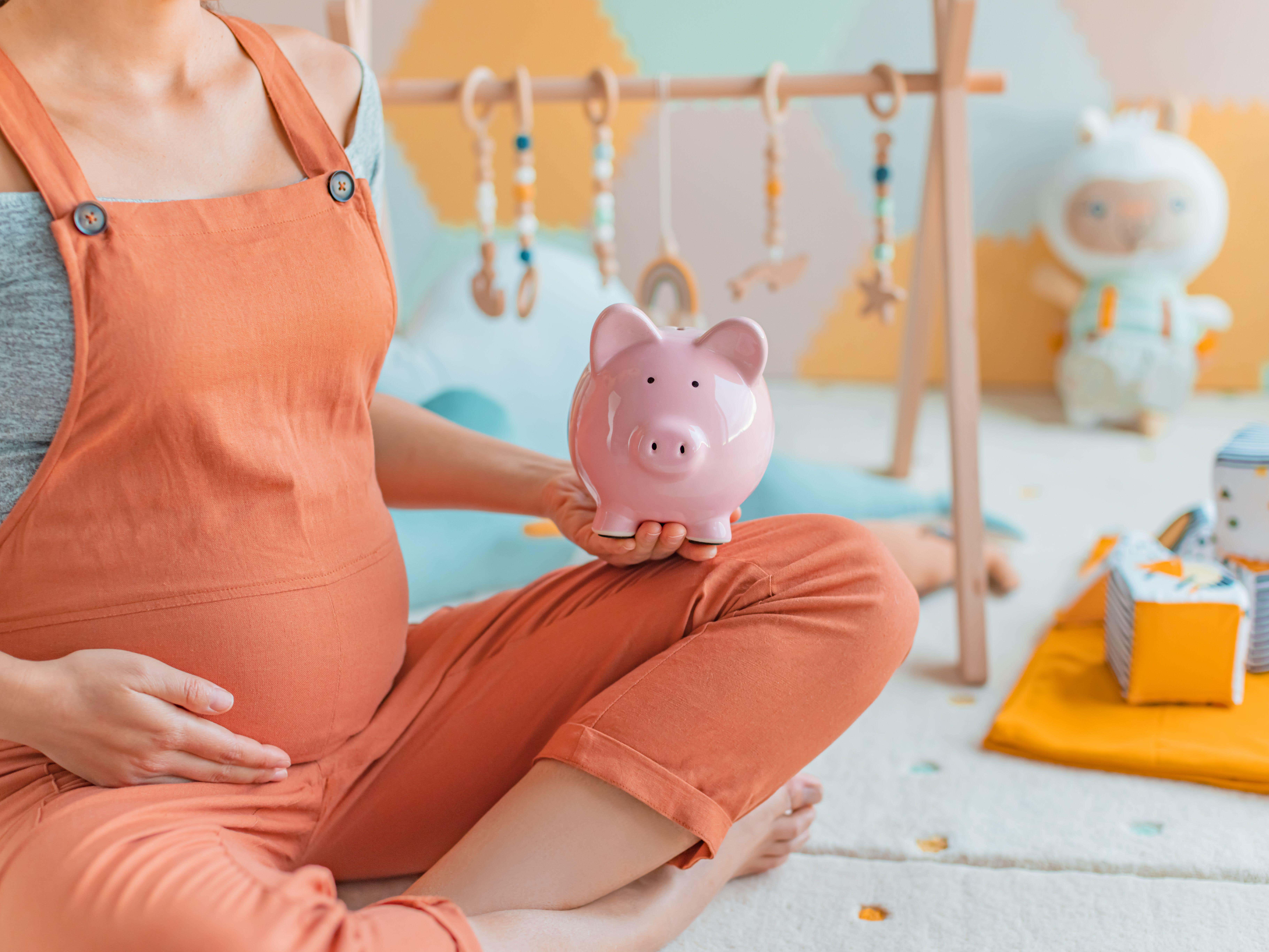 Cropped image of pregnant woman, who is wearing orange dungarees, sitting in a nursery room holding a piggy bank